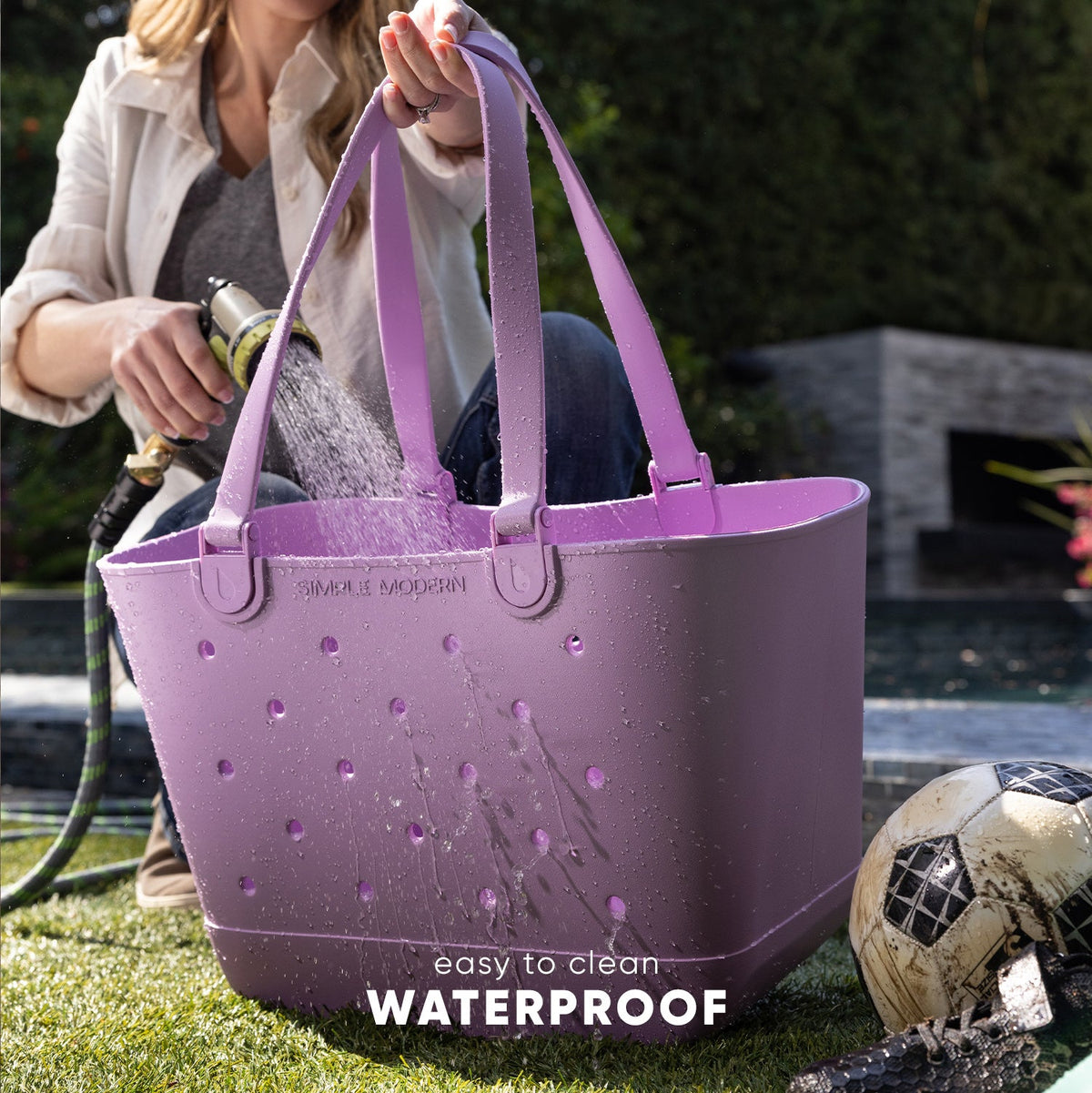 Lavender Simple Modern Getaway Tote being sprayed with water from a garden hose in a backyard to demonstrate its waterproof capability. Woman holding the bag with water droplets cascading off its ventilated surface.