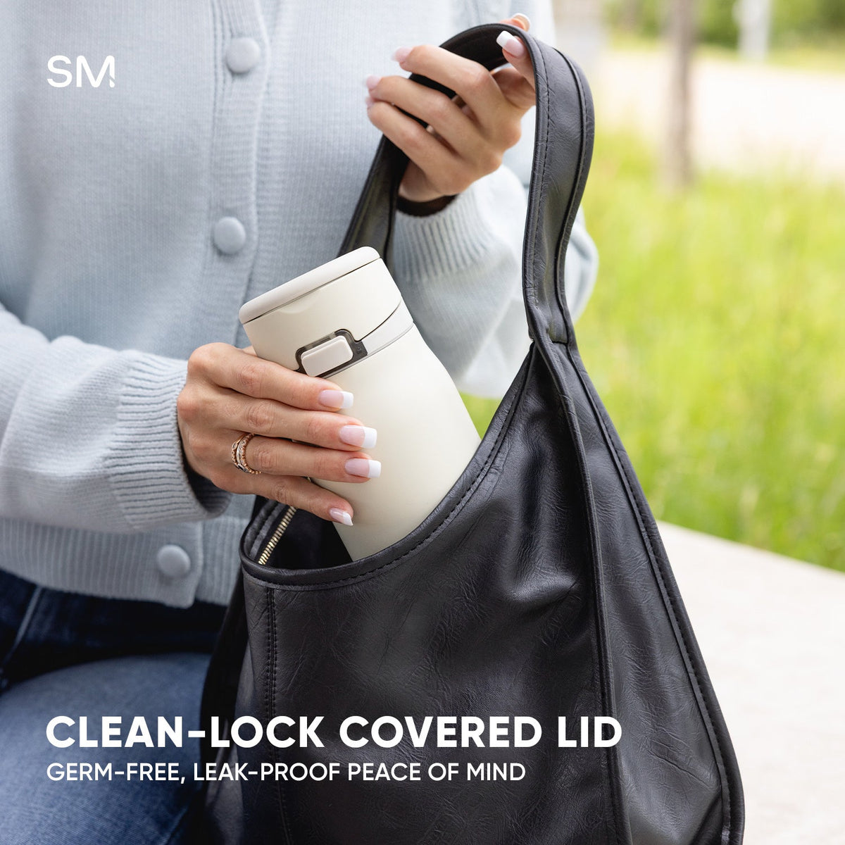 Woman holding cream-colored Simple Modern tumbler with Clean-Lock covered lid, placing it in a charcoal gray insulated carrying bag outdoors in a park setting.